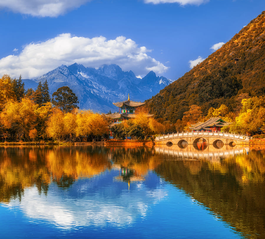 Paisaje de otoño en Yunnan con árboles de hojas amarillas reflejados en un lago cristalino, un templo clásico chino y montañas nevadas.