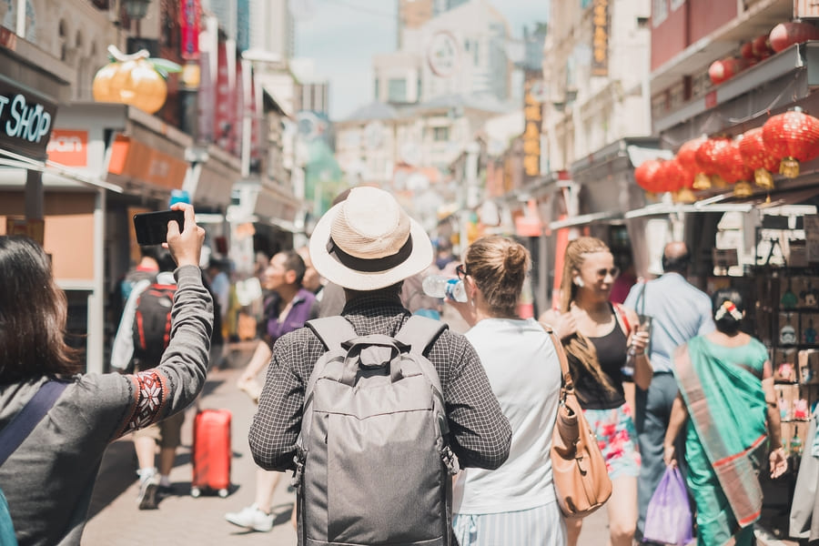 Viajero con mochila y sombrero caminando de espaldas por una concurrida calle tradicional en China decorada con linternas rojas.